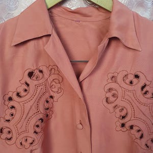 May include: A dusty rose button-down shirt featuring a pointed collar and intricate, symmetrical cut-out embroidery on both sides of the chest. The garment is displayed on a wooden hanger, showcasing a smooth fabric texture and matching buttons.