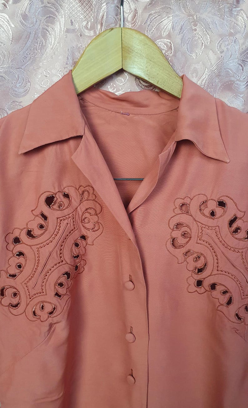 May include: A dusty rose button-down shirt featuring a pointed collar and intricate, symmetrical cut-out embroidery on both sides of the chest. The garment is displayed on a wooden hanger, showcasing a smooth fabric texture and matching buttons.