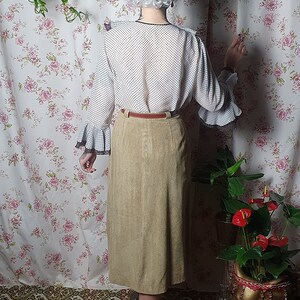 May include: A woman wearing a white blouse with black polka dots and a brown suede skirt. She is standing in front of a floral patterned wall with a plant hanging from the ceiling.