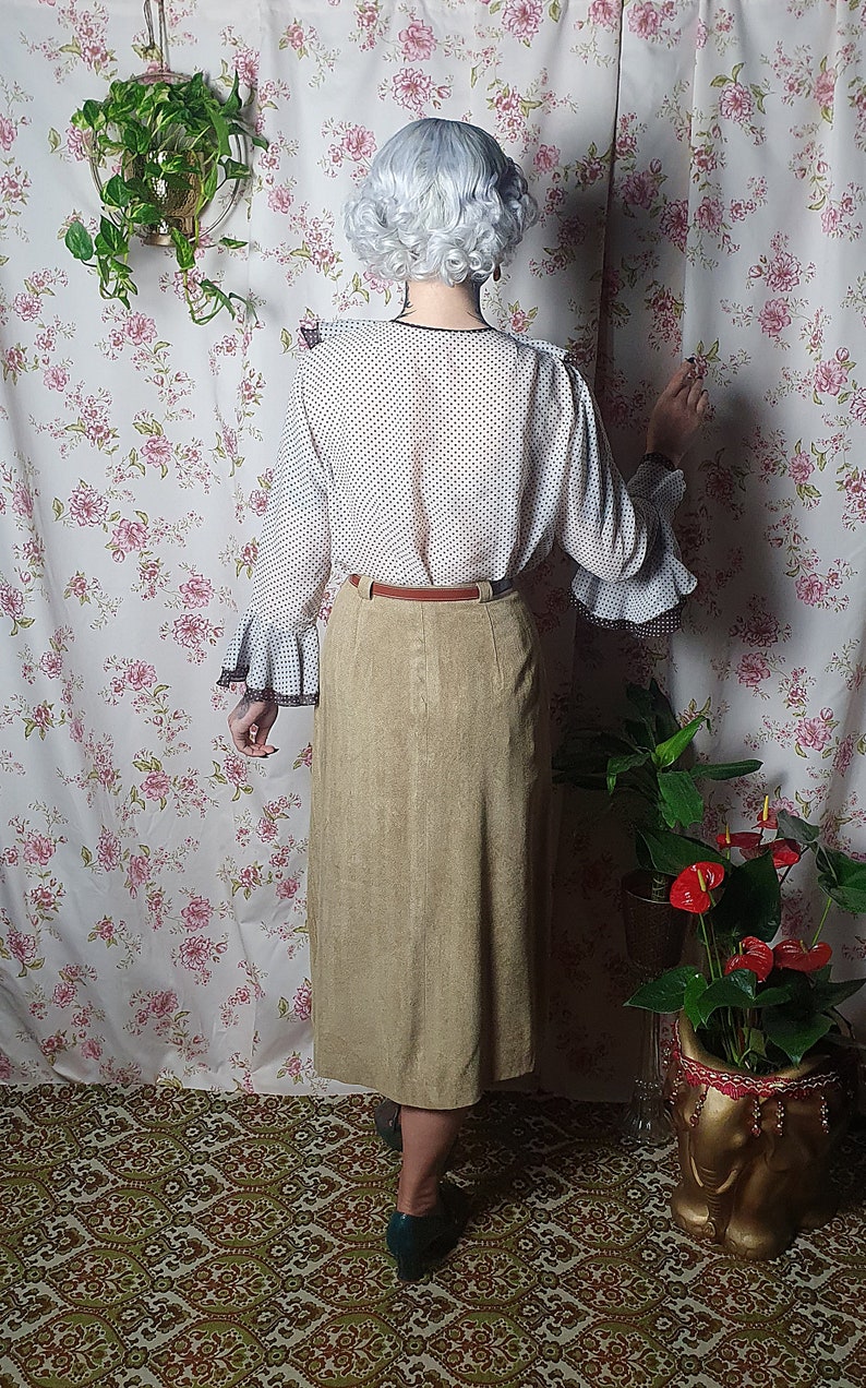May include: A woman wearing a white blouse with black polka dots and a brown suede skirt. She is standing in front of a floral patterned wall with a plant hanging from the ceiling.