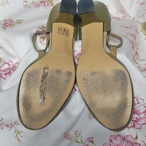 Op de afbeelding: Een paar olijfgroene sandalen met hakken en een T-band ontwerp. De zolen vertonen slijtage en de hakken zijn donker. De sandalen staan tegen een bloemig patroon.