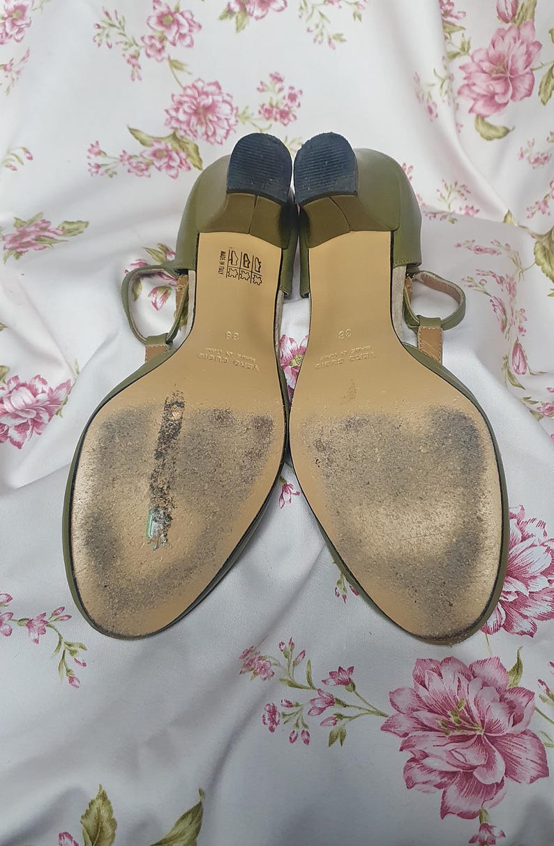 Op de afbeelding: Een paar olijfgroene sandalen met hakken en een T-band ontwerp. De zolen vertonen slijtage en de hakken zijn donker. De sandalen staan tegen een bloemig patroon.