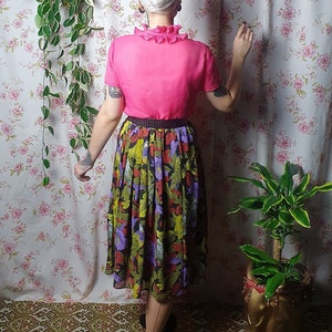 May include: A woman wearing a pink blouse with a ruffled collar and a black and green floral skirt. She is standing in front of a floral patterned wall with a hanging plant and a potted plant.