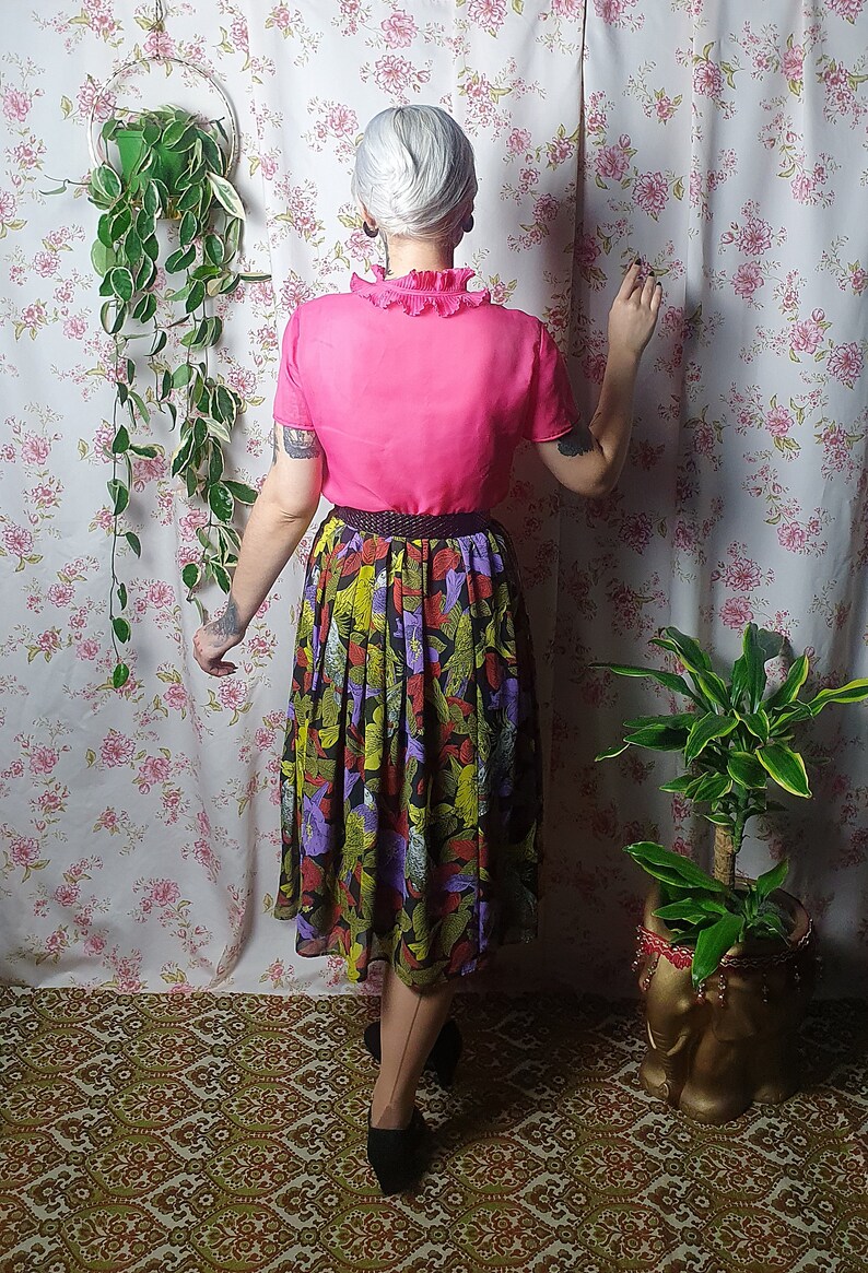May include: A woman wearing a pink blouse with a ruffled collar and a black and green floral skirt. She is standing in front of a floral patterned wall with a hanging plant and a potted plant.