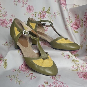 Op de afbeelding: Een paar olijfgroene en gele schoenen in vintage-stijl met een T-band ontwerp. De schoenen hebben een lage hak en een gespsluiting. De achtergrond is een stof met bloemenpatroon en roze bloemen.