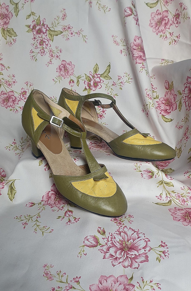Op de afbeelding: Een paar olijfgroene en gele schoenen in vintage-stijl met een T-band ontwerp. De schoenen hebben een lage hak en een gespsluiting. De achtergrond is een stof met bloemenpatroon en roze bloemen.