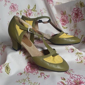 Op de afbeelding: Een paar olijfgroene en gele leren schoenen met een T-band ontwerp. De schoenen hebben een kleine hak en een gespsluiting. De schoenen worden gepresenteerd op een stof met bloemenpatroon.