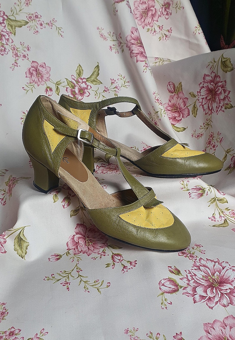 Op de afbeelding: Een paar olijfgroene en gele leren schoenen met een T-band ontwerp. De schoenen hebben een kleine hak en een gespsluiting. De schoenen worden gepresenteerd op een stof met bloemenpatroon.