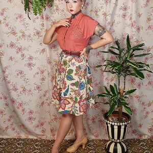 May include: A short-sleeved coral button-down shirt with decorative embroidery on the chest, paired with a cream-colored knee-length skirt featuring a multi-colored floral pattern, a thin brown belt, and beige heeled shoes with a cross-strap design.