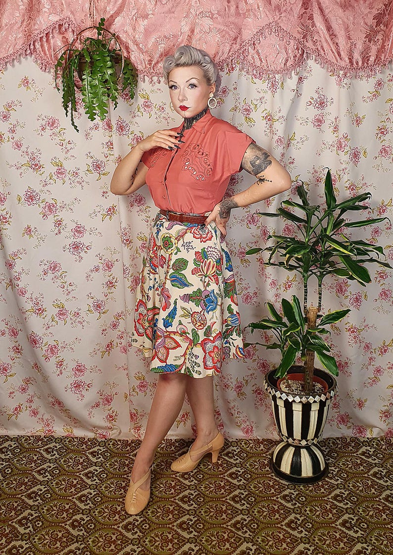 May include: A short-sleeved coral button-down shirt with decorative embroidery on the chest, paired with a cream-colored knee-length skirt featuring a multi-colored floral pattern, a thin brown belt, and beige heeled shoes with a cross-strap design.