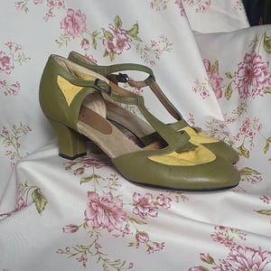 Op de afbeelding: Olijfgroene en gele schoenen in vintage-stijl met een T-strap ontwerp. De schoenen hebben een ronde neus en een kleine hak. De bovenkant is olijfgroen leer met gele accenten. De schoenen worden gepresenteerd op een stof met bloemenpatroon.