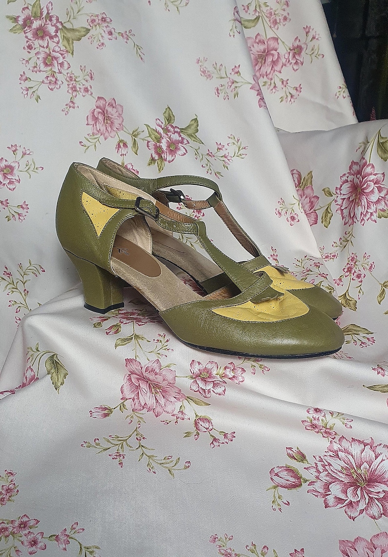 Op de afbeelding: Olijfgroene en gele schoenen in vintage-stijl met een T-strap ontwerp. De schoenen hebben een ronde neus en een kleine hak. De bovenkant is olijfgroen leer met gele accenten. De schoenen worden gepresenteerd op een stof met bloemenpatroon.