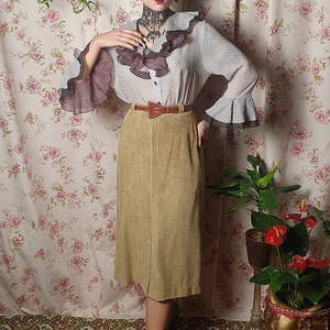 May include: A woman wearing a white and black polka dot blouse with a brown ruffle trim and a brown belt. She is wearing a brown corduroy skirt and green shoes. She is standing in front of a floral patterned wall.