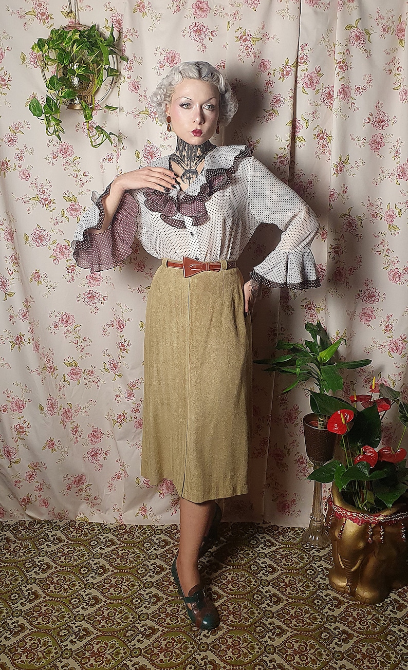 May include: A woman wearing a white and black polka dot blouse with a brown ruffle trim and a brown belt. She is wearing a brown corduroy skirt and green shoes. She is standing in front of a floral patterned wall.
