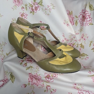 Op de afbeelding: Olijfgroene en gele vintage T-strap pumps. De schoenen hebben een ronde neus, een kleine hak en een gespsluiting. De bovenkant is olijfgroen leer met gele accenten. De schoenen worden gepresenteerd op een stof met bloemenpatroon.