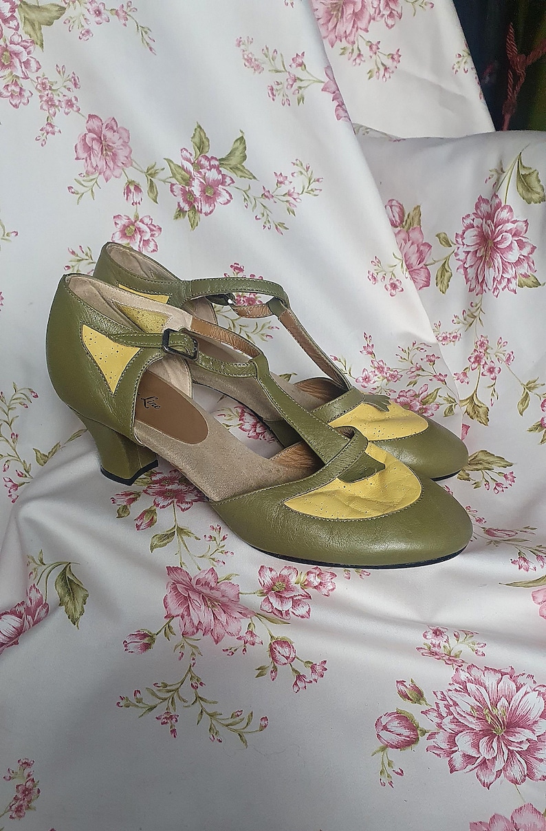 Op de afbeelding: Olijfgroene en gele vintage T-strap pumps. De schoenen hebben een ronde neus, een kleine hak en een gespsluiting. De bovenkant is olijfgroen leer met gele accenten. De schoenen worden gepresenteerd op een stof met bloemenpatroon.
