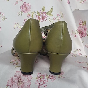 Op de afbeelding: Olijfgroene leren pumps met een T-band ontwerp. De schoenen hebben een ronde neus en een kleine, stevige hak. De achtergrond is een bloemenpatroon in roze en wit.