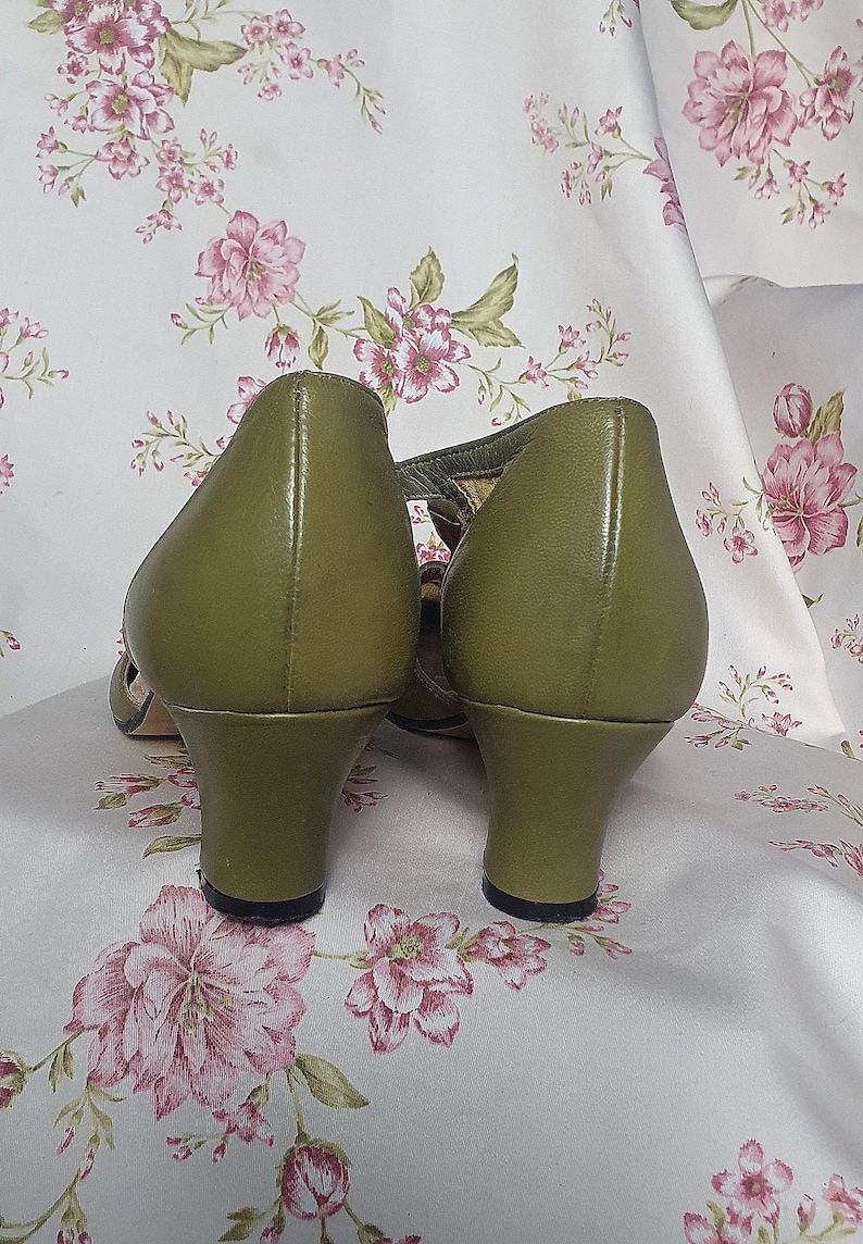 Op de afbeelding: Olijfgroene leren pumps met een T-band ontwerp. De schoenen hebben een ronde neus en een kleine, stevige hak. De achtergrond is een bloemenpatroon in roze en wit.