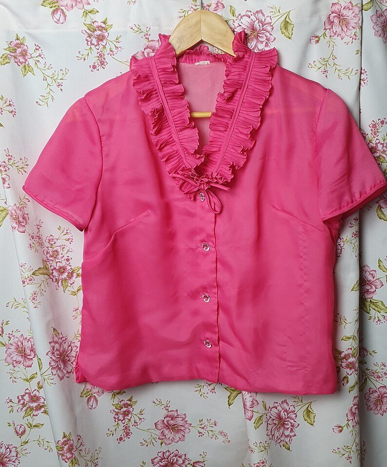 May include: A pink short-sleeved blouse with a ruffled collar and button closure.