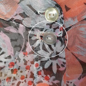 以下が含まれることがあります： 花柄のプリントが施された、透き通るようなピーチ色の生地のクローズアップ。生地には、穴に通された黒い糸で留められた白いボタンが2つ付いています。生地には「Machine Gentle Tumble」と書かれたラベルが付いています。