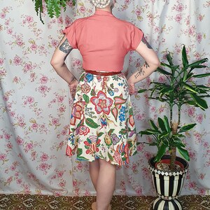 May include: A short-sleeved coral-colored blouse paired with a cream-colored skirt featuring a vibrant, multi-colored floral and paisley print. The skirt has a flared silhouette and is cinched at the waist with a thin, dark brown leather belt.