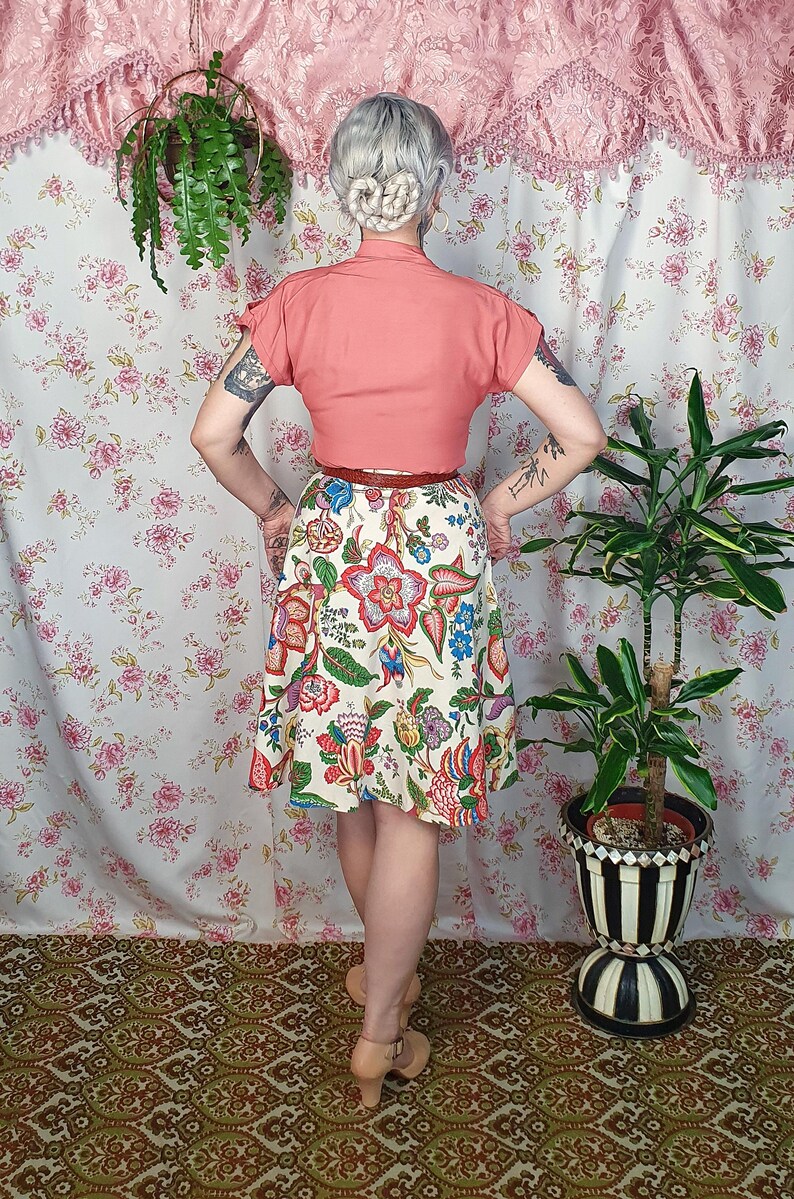 May include: A short-sleeved coral-colored blouse paired with a cream-colored skirt featuring a vibrant, multi-colored floral and paisley print. The skirt has a flared silhouette and is cinched at the waist with a thin, dark brown leather belt.
