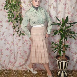Op de afbeelding: Een lichtgroen overhemd met lange mouwen, knoopsluiting, een subtiel ruitpatroon en donkere bloemaccenten, gecombineerd met een beige geplooide midirok met horizontale strepen, een witte tailleband en witte peep-toe hakken.