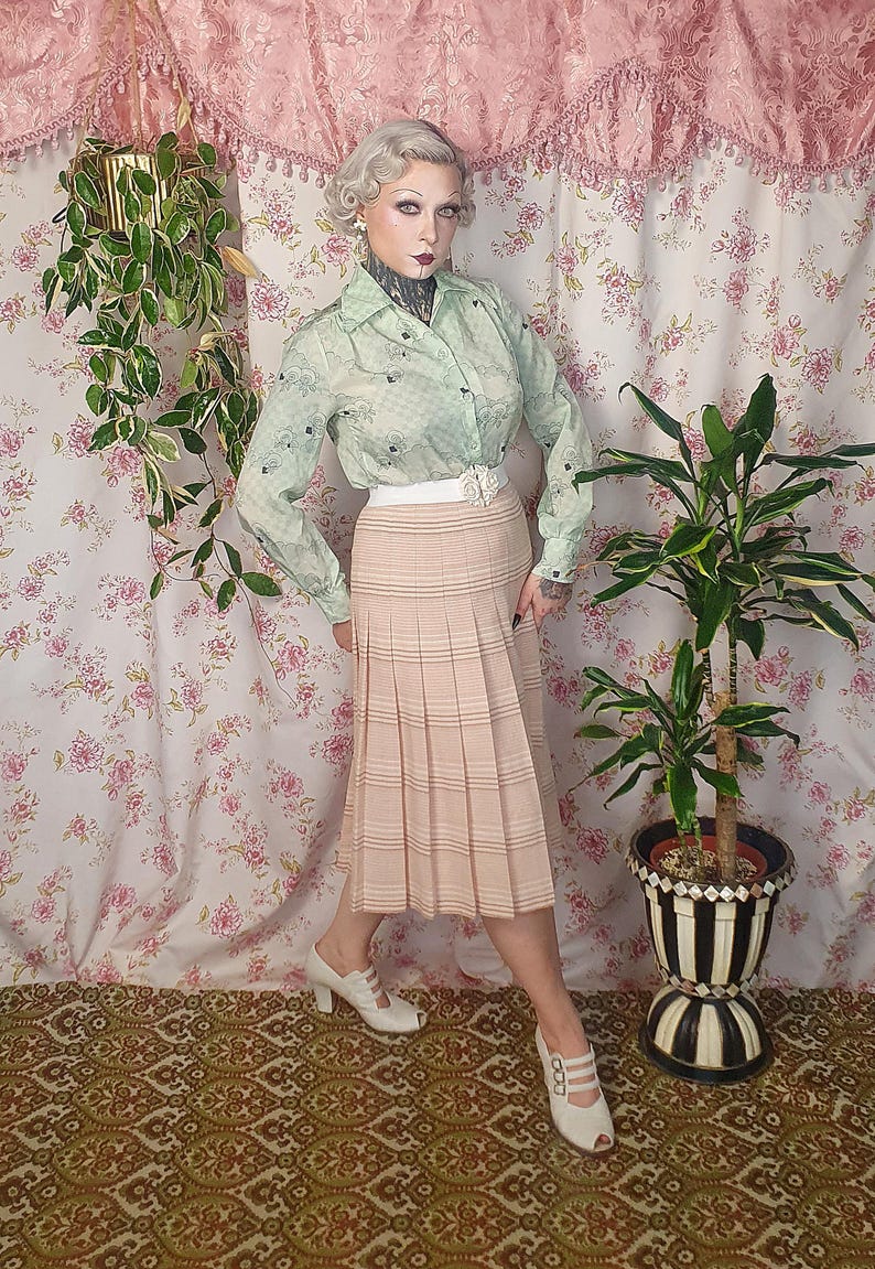 Op de afbeelding: Een lichtgroen overhemd met lange mouwen, knoopsluiting, een subtiel ruitpatroon en donkere bloemaccenten, gecombineerd met een beige geplooide midirok met horizontale strepen, een witte tailleband en witte peep-toe hakken.