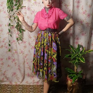 May include: A woman wearing a pink blouse with a ruffled collar and a black and green floral skirt. She is standing in front of a floral patterned wall with a hanging plant.
