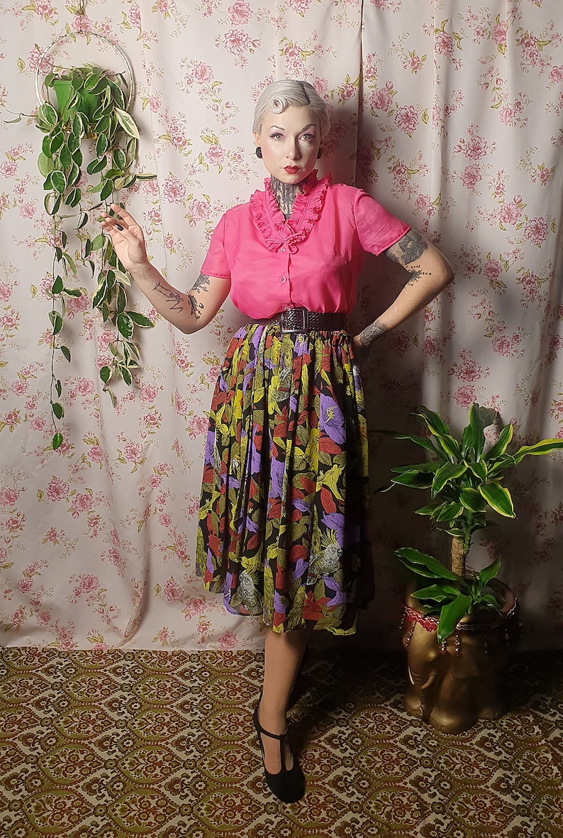 May include: A woman wearing a pink blouse with a ruffled collar and a black and green floral skirt. She is standing in front of a floral patterned wall with a hanging plant.