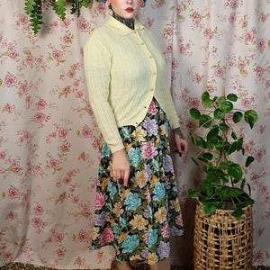 May include: A vintage-style outfit featuring a pale yellow cardigan with button closures, paired with a floral print midi skirt in black, blue, pink, and purple. The outfit is completed with brown heels. The background has a floral pattern.
