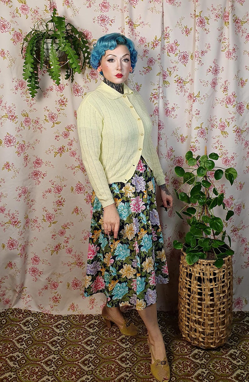 May include: A vintage-style outfit featuring a pale yellow cardigan with button closures, paired with a floral print midi skirt in black, blue, pink, and purple. The outfit is completed with brown heels. The background has a floral pattern.