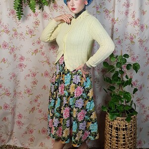 May include: A woman wearing a light yellow cardigan and a floral print skirt. The skirt features a black background with large, colorful flowers. She is wearing yellow shoes. A woven basket with a plant is to her right.
