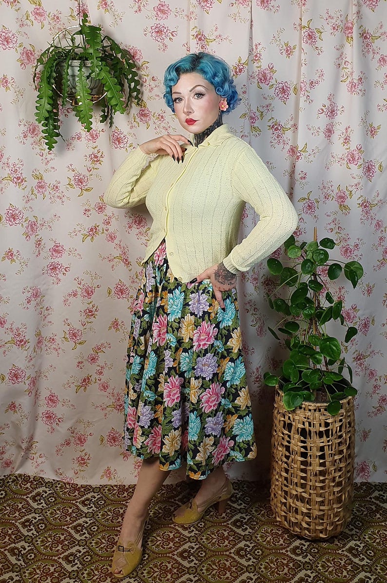 May include: A woman wearing a light yellow cardigan and a floral print skirt. The skirt features a black background with large, colorful flowers. She is wearing yellow shoes. A woven basket with a plant is to her right.