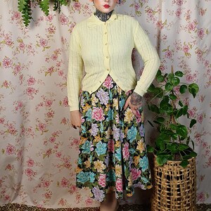 May include: A light yellow cardigan with a collared neckline and button closure. The cardigan is paired with a floral print skirt featuring pink, blue, and yellow flowers on a black background. The outfit is completed with tan shoes.