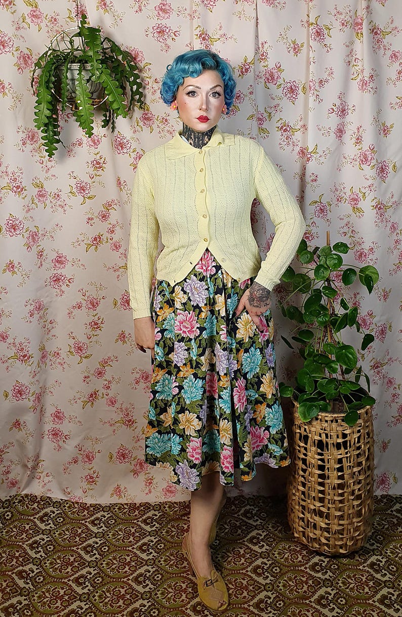 May include: A light yellow cardigan with a collared neckline and button closure. The cardigan is paired with a floral print skirt featuring pink, blue, and yellow flowers on a black background. The outfit is completed with tan shoes.