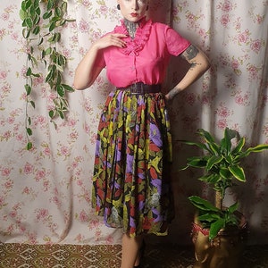 May include: A woman wearing a pink blouse with a ruffled collar and a black and green floral skirt. She is standing in front of a floral patterned wall with a plant hanging on the left and another plant in a gold pot on the right.