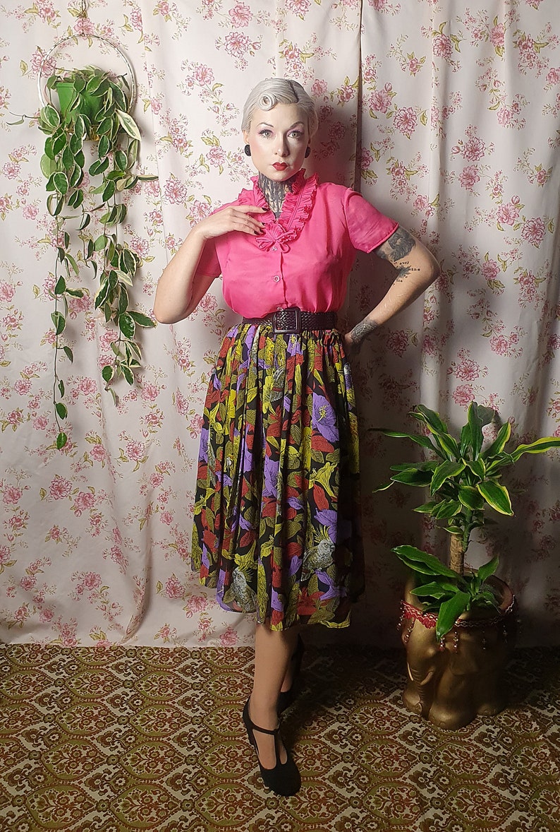 May include: A woman wearing a pink blouse with a ruffled collar and a black and green floral skirt. She is standing in front of a floral patterned wall with a plant hanging on the left and another plant in a gold pot on the right.
