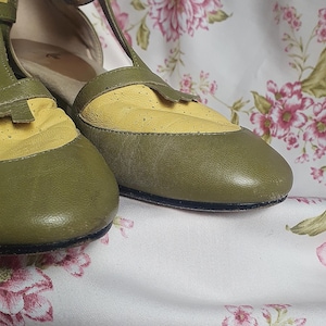 Op de afbeelding: Olijfgroene en gele leren Mary Jane schoenen. De schoenen hebben een geperforeerde gele neus en een bandje met een gespsluiting. De schoenen staan tegen een bloemenachtergrond.