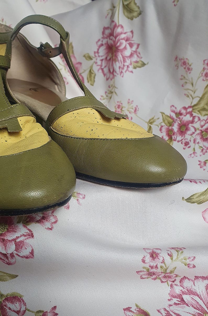 Op de afbeelding: Olijfgroene en gele leren Mary Jane schoenen. De schoenen hebben een geperforeerde gele neus en een bandje met een gespsluiting. De schoenen staan tegen een bloemenachtergrond.