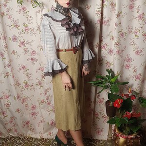 May include: A woman wearing a white and brown polka dot blouse with a brown ruffled collar and a brown belt. She is wearing a brown skirt and green shoes. She is standing in front of a floral patterned wall.