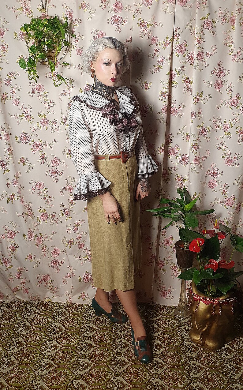 May include: A woman wearing a white and brown polka dot blouse with a brown ruffled collar and a brown belt. She is wearing a brown skirt and green shoes. She is standing in front of a floral patterned wall.