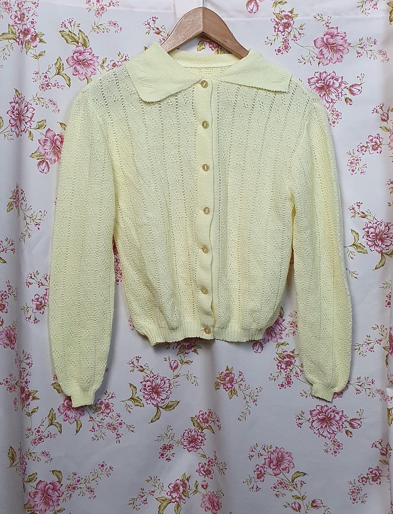 May include: A pale yellow cardigan with a rounded collar and long sleeves. The button-down sweater has a textured knit pattern and is hanging on a wooden hanger. The background is a floral patterned fabric.