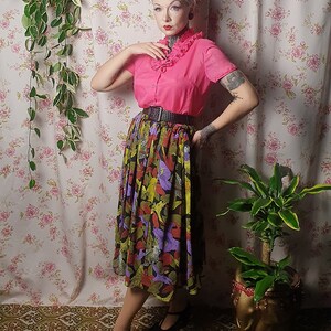 May include: A woman wearing a pink blouse with a ruffled collar, a black belt, and a colorful floral skirt. She is standing in front of a floral patterned wall with plants in pots.