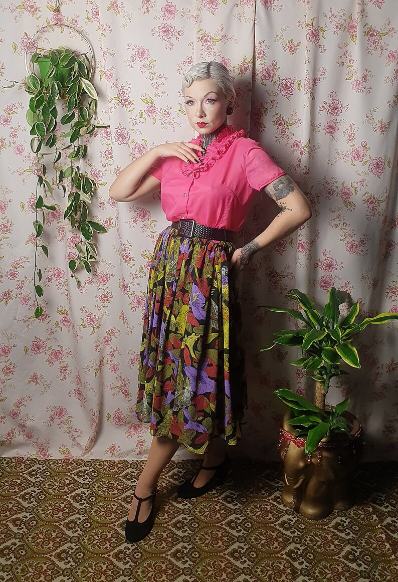 May include: A woman wearing a pink blouse with a ruffled collar, a black belt, and a colorful floral skirt. She is standing in front of a floral patterned wall with plants in pots.