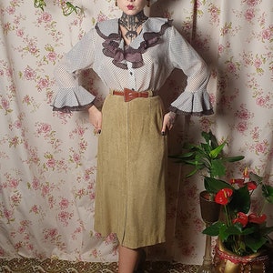 May include: A woman wearing a white blouse with black polka dots and a brown suede skirt. She is standing in front of a floral patterned wall with plants in pots.
