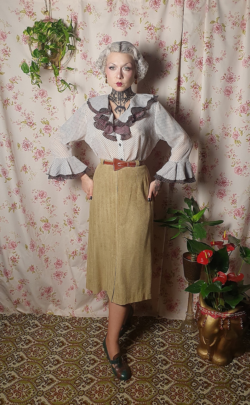 May include: A woman wearing a white blouse with black polka dots and a brown suede skirt. She is standing in front of a floral patterned wall with plants in pots.