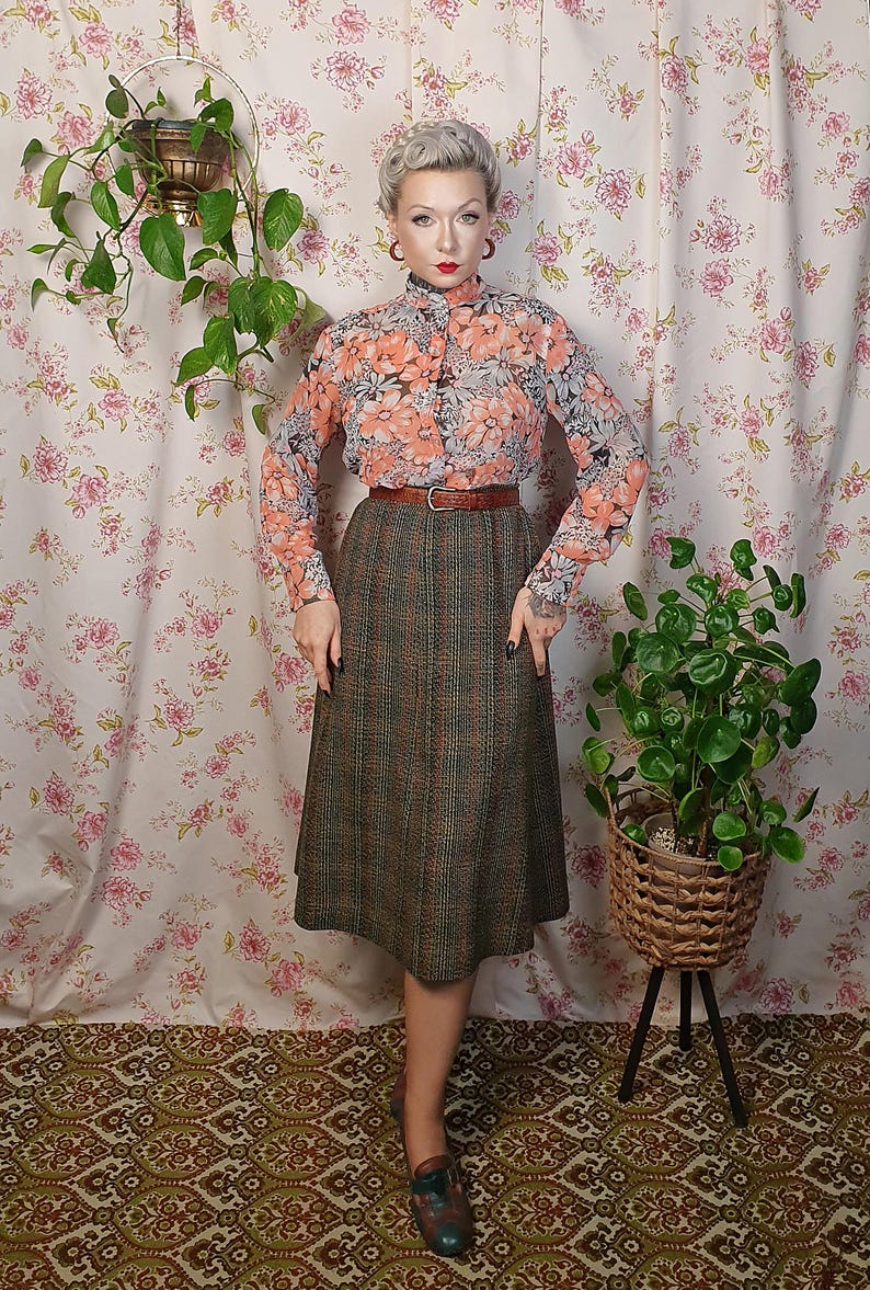 May include: A woman wearing a floral print blouse with a high collar and a brown plaid skirt. She is standing in front of a floral patterned background with a potted plant on a stand to the right.
