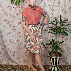 May include: A coral short-sleeved button-down shirt with decorative embroidery on the chest, paired with a cream-colored knee-length skirt featuring a vibrant multi-colored floral pattern, a thin brown belt, and tan heeled shoes.