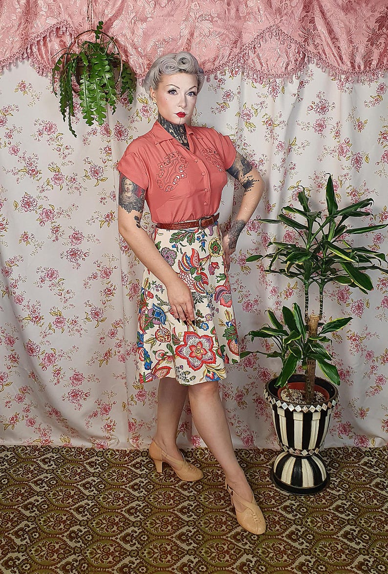 May include: A coral short-sleeved button-down shirt with decorative embroidery on the chest, paired with a cream-colored knee-length skirt featuring a vibrant multi-colored floral pattern, a thin brown belt, and tan heeled shoes.